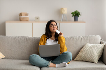 Thoughtful middle eastern lady holding credit card and using laptop, dreaming about new clothes, purchasing via internet