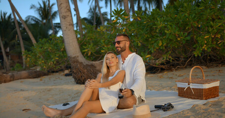 Handsome man and pretty woman sitting by the ocean on the sand beach, clinking glasses with red wine