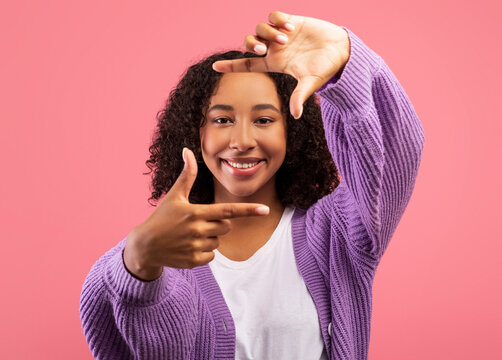 Cheerful black woman making picture frame with fingers, looking at camera and smiling for photo on pink background