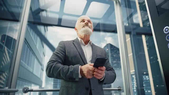 Successful Middle Aged Businessman Riding Glass Elevator to Office in Modern Business Center. Handsome Happy Man Using Smartphone, Write a Message, Social Media and Work Emails in a Lift.