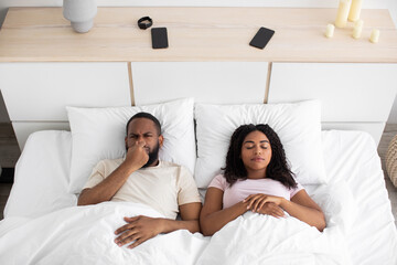 Sad disgruntled millennial african american husband closes nose from bad scent, wife sleeps on...