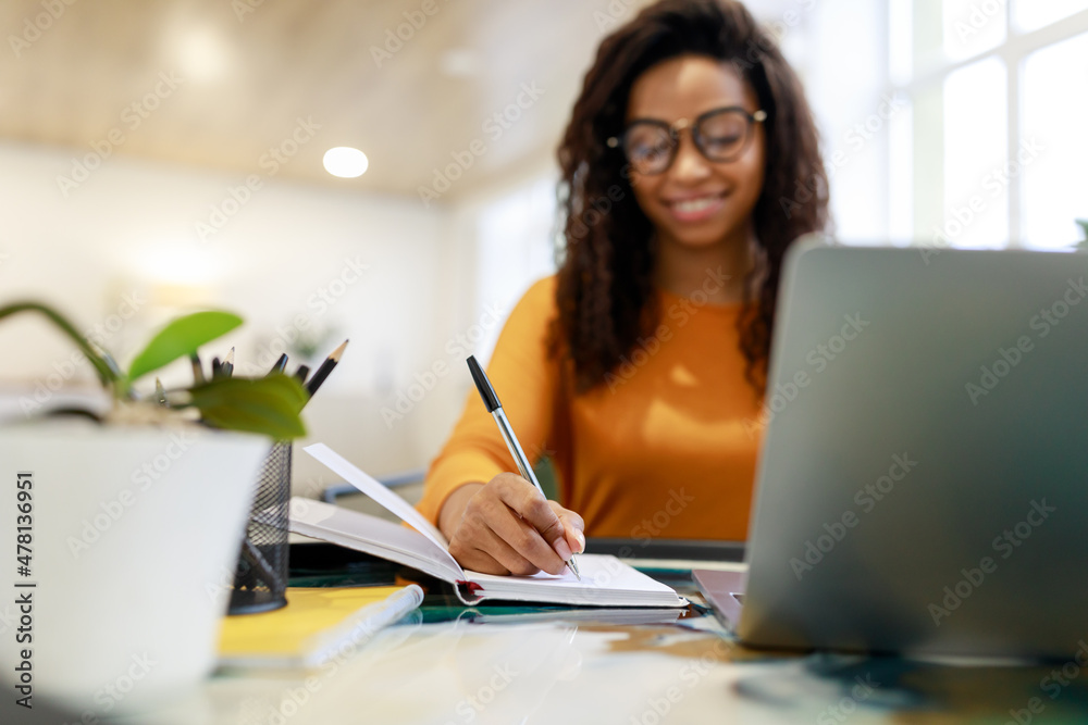 Wall mural black woman sitting at desk, using pc writing in notebook