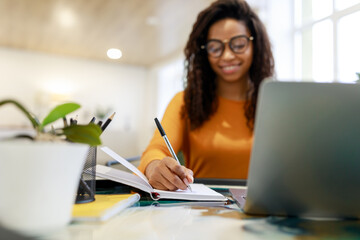 Black woman sitting at desk, using pc writing in notebook