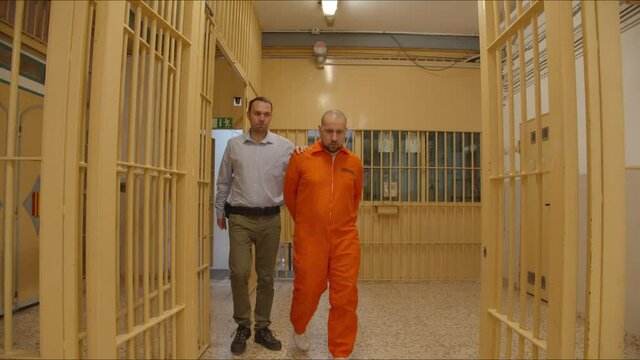 The prison guard escorts a serious prisoner in orange clothes down the corridor between the prison bars, 4k