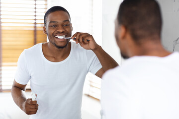 Happy Young African American Man Brushing His Teeth With Toothbrush In Bathroom
