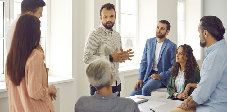 Group of people having a work meeting. Serious young male team leader, corporate manager, businessman or business coach standing in office and talking to group of employees. Website banner background