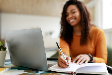 Black woman sitting at desk, using pc writing in notebook