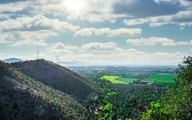 Paisaje de campos con nubes y sol sobre ellos entre monta&ntilde;as