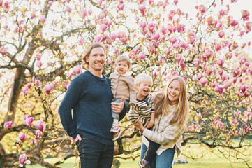 Outdoor portrait of happy young family playing in spring park under blooming magnolia tree, lovely couple with two little children having fun in sunny garden © annanahabed