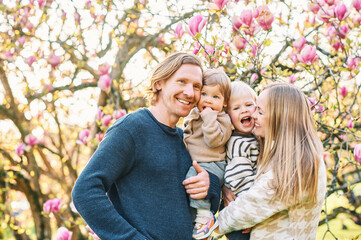 Outdoor portrait of happy young family playing in spring park under blooming magnolia tree, lovely...
