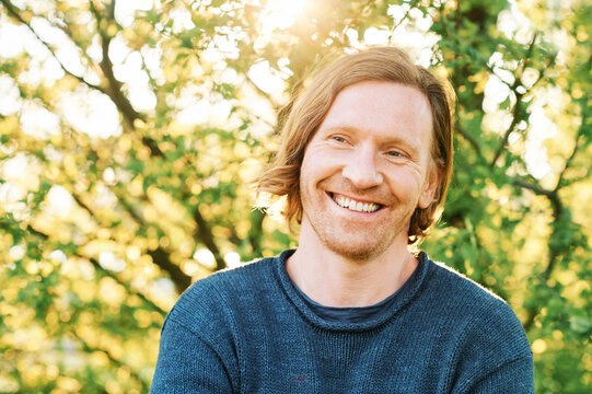 Outdoor portrait of handsome 35 - 40 year old man with red hair, posing in green sunny park, wearing blue pullover