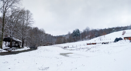 Fototapeta na wymiar Hilly countryside in winter, empty animal pen against forest and stream, land covered with snow