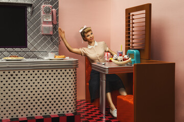Young blonde woman looking like a retro doll, staged as a customer waving at someone, in a toy reproducing a vintage dinner restaurant