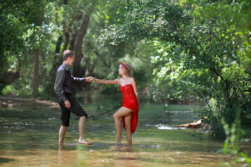 A guy and a beautiful young blonde girl with long hair in a red dress are having fun holding hands in the river water.