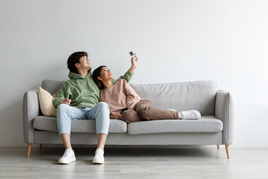 Split System. Happy Young Asian Couple With Remote Relaxing Under Air Conditioner, Sitting On Couch At Home, Mockup