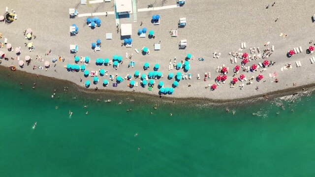 Aerial view of sea waves, umbrellas, green palms on the beach with pebbles at sunset. Summer in Batumi, Georgia. Tropical landscape with palms, people, umbrellas, sand, blue water. Aerial top view