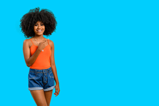 A smiling young woman with dark skin and afro hairstyle points her finger at a space on the right side of the image, she is wearing an orange bustier and blue denim shorts,  on a blue background