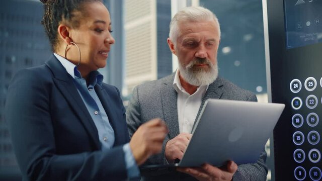 Middle Aged Businessman Talking with International Investment Partner while Riding Glass Elevator to Office in a Modern Business Center. Corporate Associates Use Laptop Computer in a Lift.