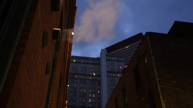 An evening view of steam exiting a pipe on a building in the alley of a large city.  	