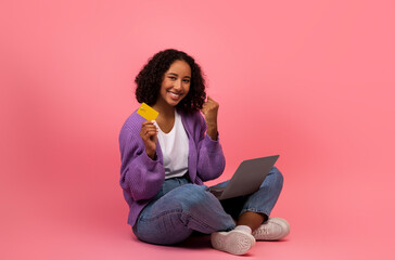 Young black woman with credit card and laptop making YES gesture, excited over big sale in online store, winning lottery