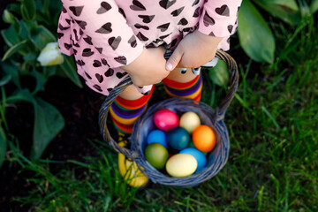 Close-up of legs of toddler girl with colorful stockings and shoes and basket with colored eggs. Child having fun with traditional Easter eggs hunt, outdoors. Celebration of christian holiday.