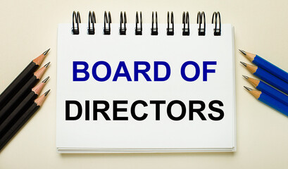 On a light background, a white notebook with the text BOARD OF DIRECTORS and black and blue pencils...