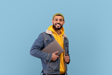 Naklejka premium Happy middle eastern guy holding laptop and smiling at camera, arabic man with pc computer over blue background
