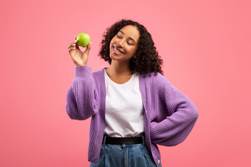 Portrait of cheerful young black woman holding ripe green apple and smiling on pink studio...