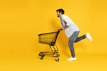 Young Caucasian man running and pushing an empty shopping cart or shopping trolley isolated on yellow background © comzeal