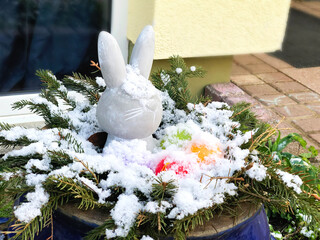 Easter outdoors decoration covered with snow. Easter bunny and colored eggs in a big bowl outside. Cold and late spring, snowfall in April.