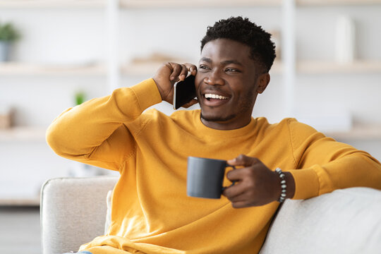 Cheerful black guy having phone conversation with friend, drinking tea