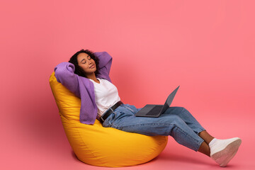 Young black woman with laptop taking break, sitting in bean bag chair with closed eyes, relaxing...