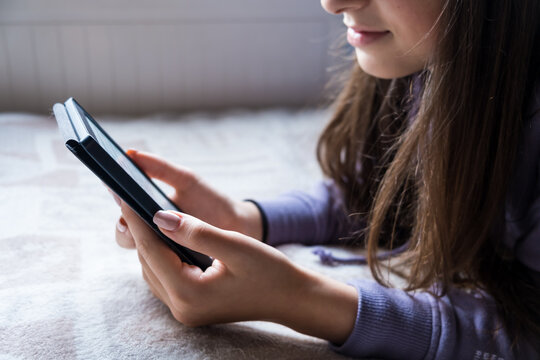 Young Teenager GIrl is focused reading an electronic romance with her Ebook Reader while she is on the bed. The text on the ebook reader is a "lorem ipsum"
