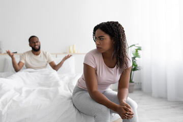 Angry millennial african american husband freaking out, swears and yells at sad unhappy wife on bed