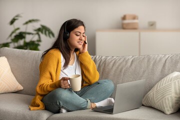 Telecommuting concept. Arab woman sitting on sofa and working online on laptop, wearing headset and holding cup