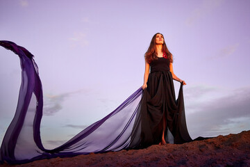 Beautiful girl with black dress and violet fluttering cloth dancing on the sand dune. Model or dancer posing in nature landscape