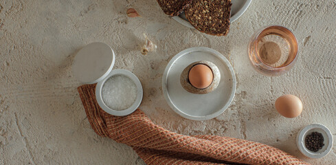 Breakfast setting with boiled egg in stoneware egg cup, whole grain rye bread, glass of water, salt flakes and pepper in concrete bowls on rough textured clay background. Healthy breakfast concept