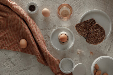 Fototapeta na wymiar Breakfast setting with boiled egg in stoneware egg cup, whole grain rye bread, glass of water, salt flakes and pepper in concrete bowls on rough textured clay background. Healthy breakfast concept