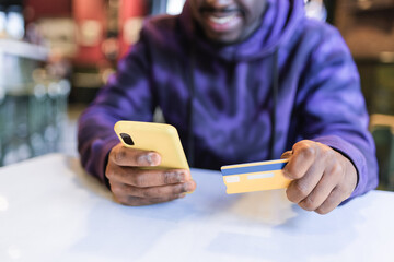Crop positive African American male client sitting at table in cafe and using cellphone for online shopping and paying with plastic card