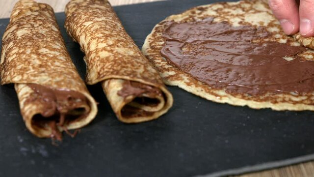 person rolling crepes spread with hazelnut chocolate cream placed on a square plate of black slate, with two more ready-made crepes on the left, 4k resolution