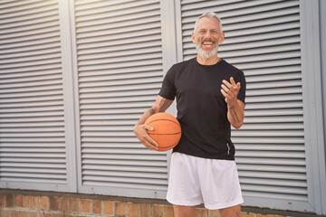 Joyful middle aged man in sportswear holding basketball ball and smiling at camera while standing...