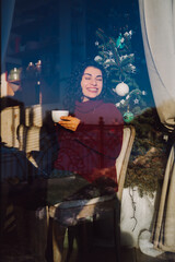 Young happy smiling curly woman drink tea or coffee in city cafe and look out the window with decorated Christmas tree in background.