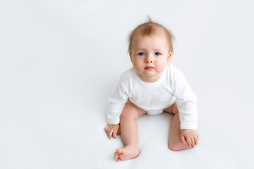 Infant with protruding hair is sitting looking at the camera. Funny baby on a white background in a bodysuit. Care and care of children from birth. The girl holds her leg for balance