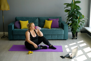 An overweight woman drinks water from a bottle sitting on the floor on a mat during an online home sports workout. Curvy young female doing pilates for weight loss