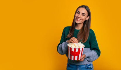 Photo of young beautiful happy woman in sweater while she holds big bucket of popcorn for watching movie in cinema and looking on camera with smile