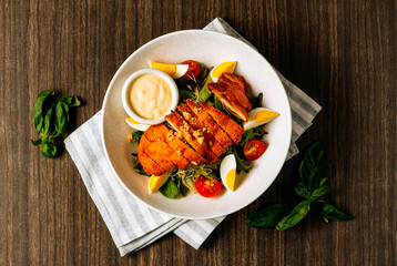 Chicken schnitzel served with fresh salad, boiled egg, tomato and mayo in white bowl on dark wood...
