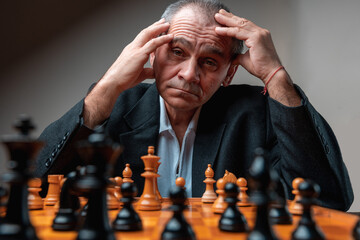 Portrait of thoughtful man concentrated on opponent on chess match, holding head with his hands. 
Classical formal suit, vintage look. Professional competition, thinking of strategy.Looking at camera.