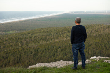 Thoughtful man with hands in his pockets, standing with his back to the camera on top of mountain,  staring at beautiful nature landscape. Outdoor forest and ocean background. copyspace. 