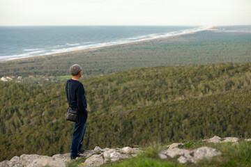 Thoughtful man with hands in his pockets,  standing on top of mountain,  staring at beautiful nature landscape. Forest and ocean background. copyspace. Inspiring and motivational view.
