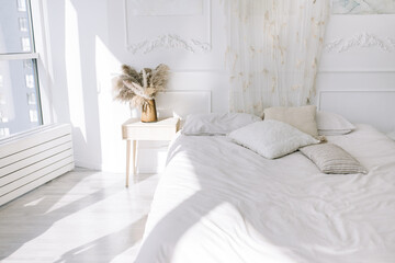 Light cozy bedroom, bed with white sheets and pillows, vase with dried flowers  near the bed. The sun shines through the window and falls into the bedroom